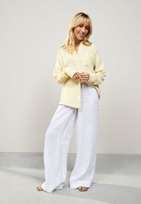 Light yellow, textured fabric button-up shirt with a chest pocket; paired with loose, white linen trousers and sandals on a neutral background.
