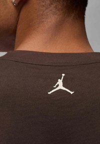 Camiseta marrón con textura acanalada, que presenta un logo Jumpman blanco en la parte superior de la espalda. La tela parece suave y con un cuello ajustado.