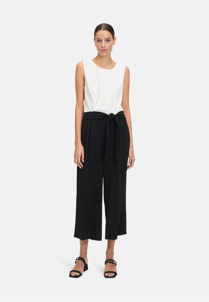 Biely top bez rukávov s voľným výstrihom, kombinovaný s čiernymi culottes s vysokým pásom a viazaným opaskom. Čierne ploché sandále dopĺňajú vzhľad.