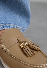 Lichtbruin leren loafers met kwastjes en gouden hardware, met contrasterende witte stiksels op de bovenkant. Buitenrand gedeeltelijk zichtbaar.