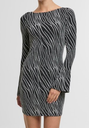 Vestido mini negro con mangas largas y patrón de rayas onduladas plateadas, silueta ajustada, llevado por una persona de pie con una mano apoyada en el muslo.