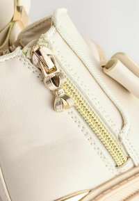 Weiße Kunstleder-Sneaker mit goldenem Reißverschluss, strukturiertem Obermaterial und runden Schnürsenkeln, ergänzt durch weitere goldene Hardware-Details.