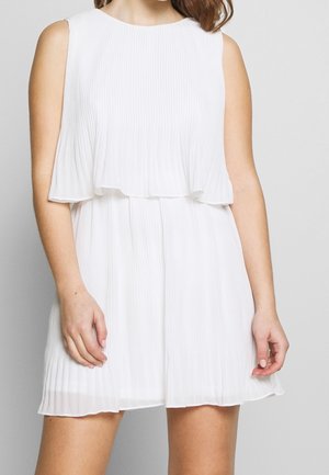 Femme portant une mini robe blanche plissée sans manches avec une jupe à couches sur un fond clair uni
