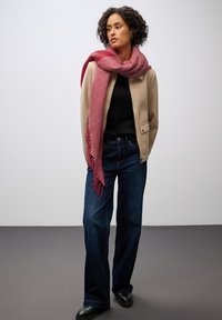 Giacca beige sopra un top nero, abbinata a jeans scuri a gamba larga. Una sciarpa rosa con frange si avvolge attorno al collo, aggiungendo texture e colore.