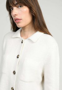 Gros plan sur une femme aux cheveux bruns portant un gilet blanc à boutons avec un col, sur un fond neutre.