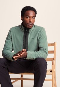 Fleece jacket in soft green with a black front zipper. Features a high collar and logo detail. Model sits on a wooden chair.