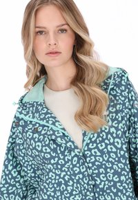 Chaqueta impermeable con base de color verde azulado oscuro y manchas de leopardo en tonos menta claro, capucha ajustable y cremallera frontal con detalles de botones.