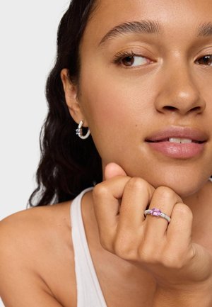 Anello d'argento con una pietra preziosa rettangolare rosa. La modella indossa un top bianco e un orecchino a cerchio con un accento blu. Texture della pelle liscia.