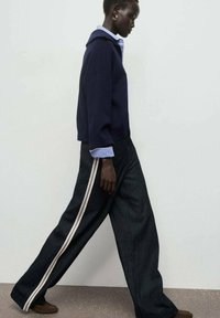Sudadera azul marino con un corte relajado, combinada con pantalones de denim oscuros de pierna ancha que tienen rayas laterales de color rosa pálido. El modelo posa de perfil.
