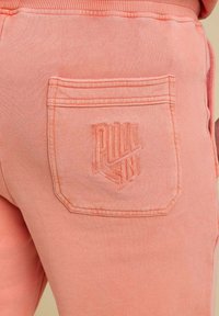 Pantalons de survêtement couleur corail avec une poche arrière et un détail de logo cousu, fabriqués en coton doux avec une texture lisse.