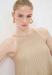 Beige geribbelde gebreide top met een hoge hals. Heeft een gouden kettingaccent langs de schouder. Model draagt grote gouden hoepeloorbellen.