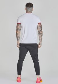Witte kortomslagen shirt met rode rand, gecombineerd met zwarte taperende broek en felroze sneakers. Zichtbare tatoeages op de armen.