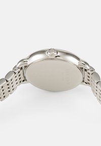 Silverfärgad metallklocka med en slät, rund urtavla och ett texturerat meshband. Har en framträdande krona och ingraverad branding på baksidan.