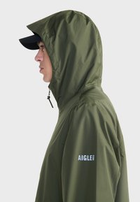 Veste imperméable vert olive avec capuche, dotée d'une fermeture éclair sur le devant, de poignets élastiques et du logo blanc "AIGLE 1853" sur le côté.