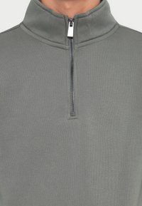Grijze hoogsluitende pullover van zacht materiaal, met een ritsluiting bij de nek. Soepele textuur met een eenvoudig, minimalistisch ontwerp.