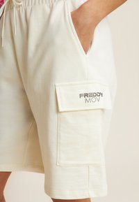 Shorts en coton de couleur crème avec une taille élastique, des poches latérales et une poche de patch proéminente arborant la marque "FREDDY MOV".