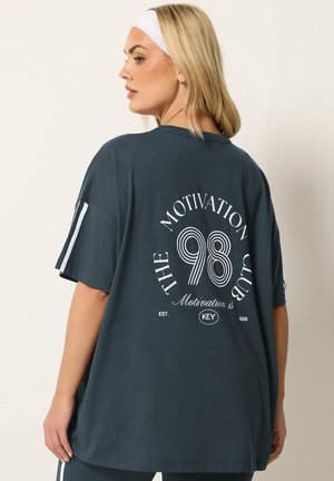 Lyshåret kvinde iført en mørk oversized t-shirt med hvid tekst "The Motivation Club 98" på ryggen og et hvidt pandebånd, set fra siden.