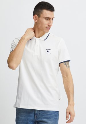 Vīrietis ar īsiem tumšiem matiem, tērpies baltā polo krekliņā ar tumši zilu apmali un logo, zilās džinsās, rāda tetovējumu uz kreisās rokas.