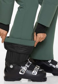 Ski pants in dark green fabric with black elastic cuffs, showcasing smooth texture and adjustable features, worn with black ski boots.