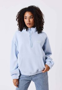 Haut de sweat à capuche bleu clair avec col zippé, poignets côtelés et grande poche frontale. Comprend des coutures contrastantes et un emblème sur la poitrine.