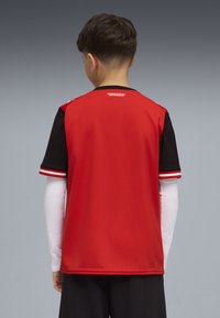 Αθλητική φανέλα κόκκινη και μαύρη με κοντά μανίκια. Κόκκινο κύριο σώμα, μαύρα μανίκια και λευκές ρίγες κατά μήκος των άκρων των μανικιών. Ελαφρύ, υφασμάτινο με υφή.