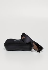Gafas de sol negras rectangulares apoyadas sobre un estuche negro con cremallera cerrado que tiene el logo "vogue" tanto en el estuche como en el marco de las gafas.