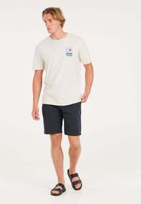 Camiseta de manga corta color beige claro con un gráfico rectangular de olas y sol, combinada con pantalones cortos azul marino y sandalias negras. Diseño casual.