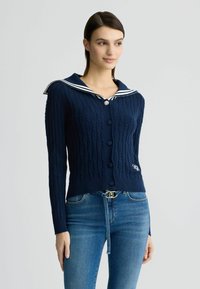 Cardigan blu navy a maglia cable con un colletto da marinaio a strisce bianche, sei bottoni neri e un dettaglio del logo sul lato sinistro in basso.