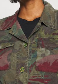 Kurtka w kamuflażu w odcieniach zieleni, czerwieni i brązu. Posiada dwie kieszenie na piersi oraz unikalne zielone guziki o geometrycznym wzorze.