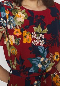 Blommig klänning med röd bakgrund, prydd med gula, blå och vita blommor. Korta ärmar och ett matchande midjeband.