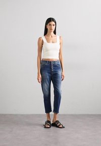 Bijela krojena crop majica, traperice s visokim strukom u plavoj boji s gumbom, i crne ravne sandale. Model stoji ispred neutralne pozadine.