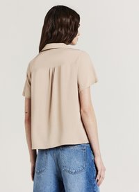Chemise beige à manches courtes avec col, dotée d'un dos plissé et d'une coupe décontractée, associée à un short en denim bleu clair.