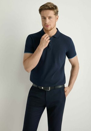 Młody mężczyzna o jasnobrązowych włosach, ubrany w granatową koszulkę polo i spodnie, poprawiający kołnierzyk jedną ręką, neutralne tło.