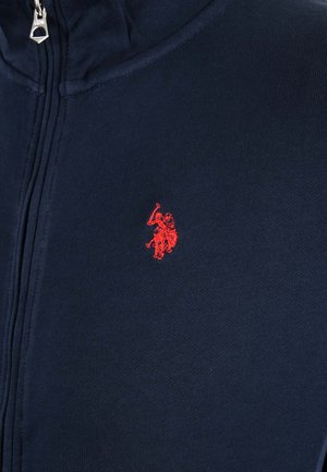 Marineblaues Kleidungsstück mit Reißverschluss und rotem, gesticktem Logo einer Person, die auf einem Pferd reitet und einen Arm hebt, der ein Objekt hält.