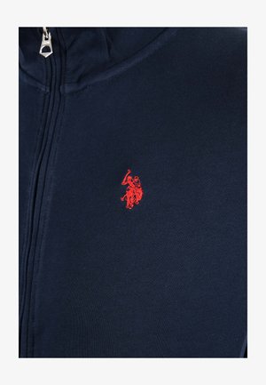 Marineblaues Kleidungsstück mit Reißverschluss und rotem, gesticktem Logo einer Person, die auf einem Pferd reitet und einen Arm hebt, der ein Objekt hält.