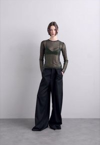 Schwarze, weit geschnittene Hosen kombiniert mit einem enganliegenden, transparenten Langarmshirt mit grünen Punkte-Mustern. Das Model steht in einer neutralen Umgebung.