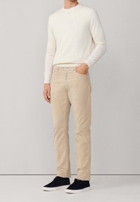 Homme portant un pull crème à manches longues, pantalon beige avec la main dans la poche, et des baskets noires, debout devant un fond uni clair.