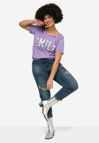 Camiseta morada con el texto "SONRÍE", jeans azules con perlas decorativas y detalles rasgados, y botas metálicas plateadas hasta la rodilla.