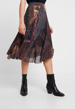 Polvipituinen kuvioitu hame, jossa on tilkkutöitä, ja mustat nilkkurit korolla henkilöllä, joka seisoo yksiväristä taustaa vasten.