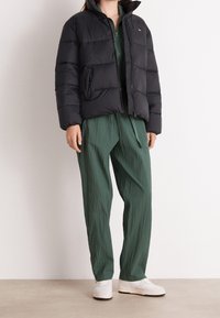 Black puffer jacket with a zip front and side pockets, paired with textured green trousers. White sneakers complete the look.
