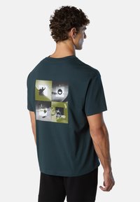 Uomo di spalle che indossa una maglietta verde scuro con quattro pannelli grafici in verde e nero sulla schiena, che presentano forme astratte e silhouette.