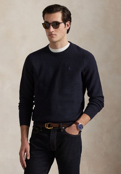 Man met donkere zonnebril, een navy trui over een wit overhemd, donkere jeans, bruine riem en polshorloge, staand met één hand in de broekzak.