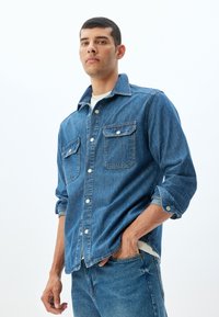Camisa de mezclilla azul con botones y cuello clásico, dos bolsillos frontales y mangas arremangadas. Lleva una camiseta blanca debajo y se combina con jeans azules.