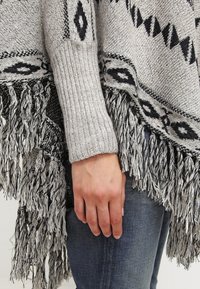 Dłoń oparta obok nogi w ciemnych dżinsach i szarym swetrze z dzianiny w czarne geometricalne wzory i frędzlami.