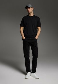 Czarny polo z subtelnym logo, w połączeniu z czarnymi dżinsami. Model nosi szary kapelusz i białe trampki, stojąc na szarym tle.