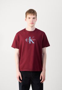 Bordo pamučna majica s kratkim rukavima i okruglim izrezom, s plavim "CK" logotipom i bijelim natpisom "Calvin Klein" ispod.