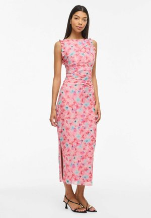 Femme portant une robe midi rose à fleurs sans manches avec une fente latérale et des détails froncés, associée à des sandales à talons noires à brides.