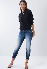 Camisa negra de botones, jeans ajustados azul con un lavado desgastado y dobladillos deshilachados en los tobillos, combinados con tacones negros con tiras.