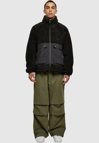 Giacca in pile nera con un pannello frontale in nylon grigio, chiusura con zip e due tasche a pattina. Abbinata a pantaloni baggy verde oliva e sneakers bianche.