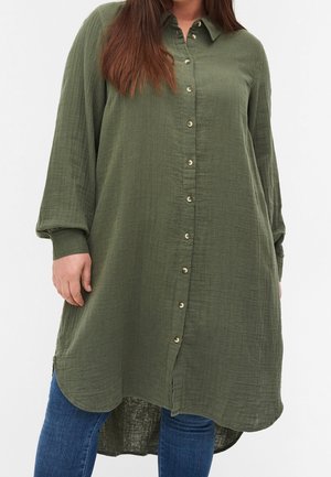 Olijfgroene blousejurk met lange mouwen en knopen, met kraag, gedragen over een blauwe spijkerbroek, waarbij de taille en bovenbenen zichtbaar zijn.